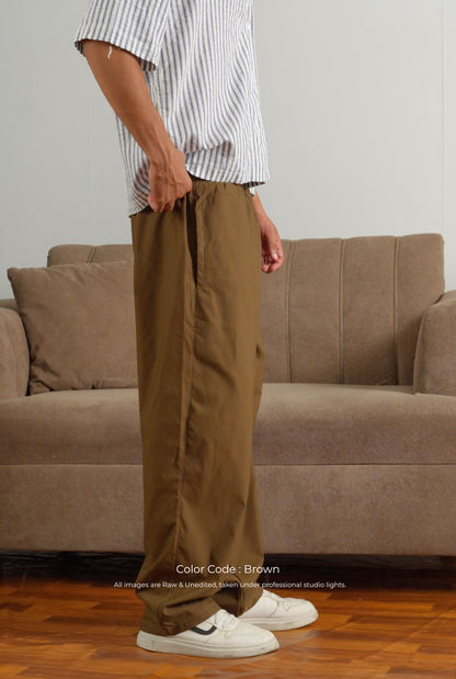 A person wearing loose brown pants, a white and navy striped short-sleeve shirt, and white sneakers, standing in front of a beige sofa in a room with a wood floor. Text at the bottom reads 'Color Code - Brown'.