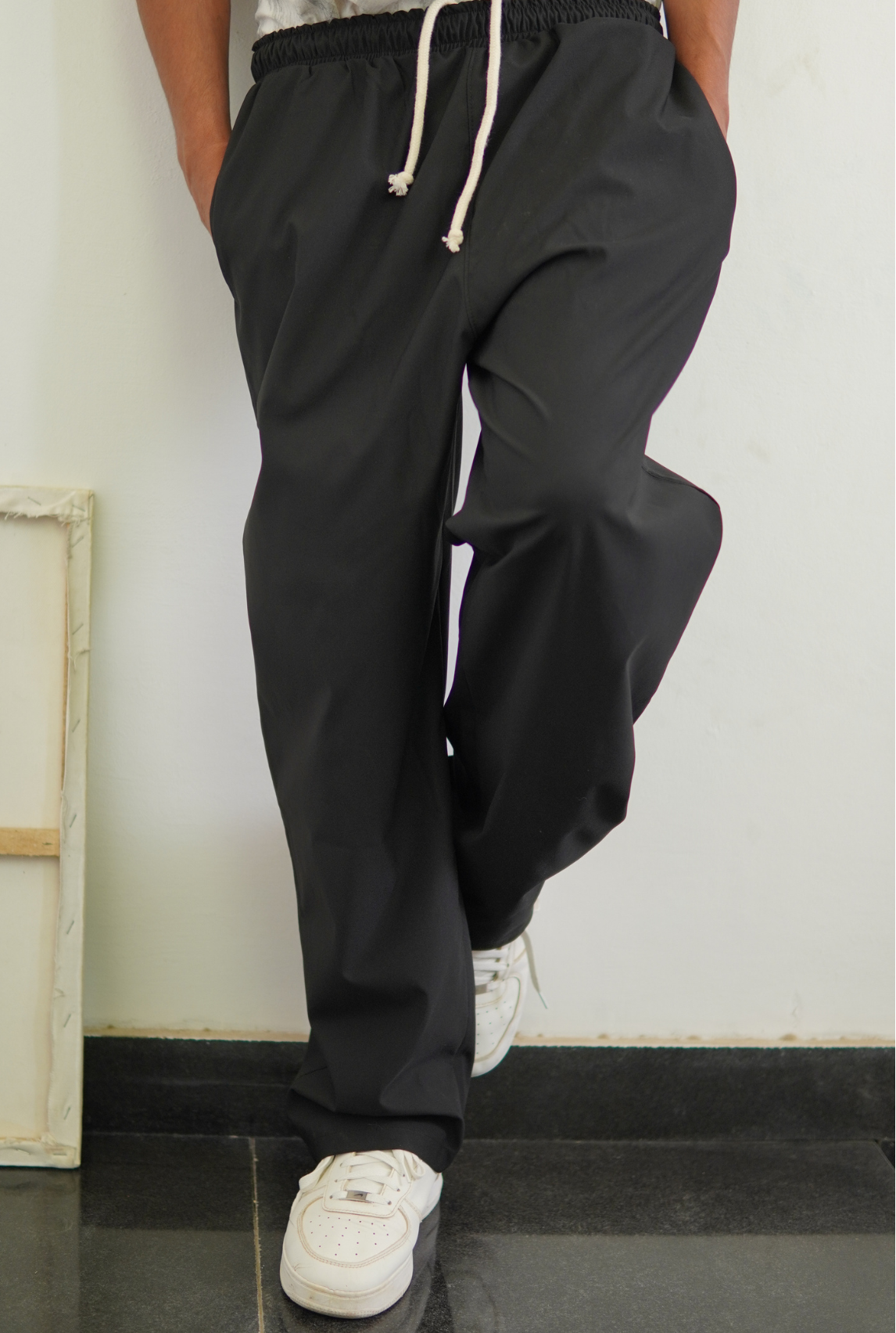 Person wearing black pants with a drawstring and white sneakers against a plain wall.